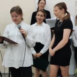 Uczennica śpiewa piosenkę, za nia stoją koleżanki i nauczycielka.